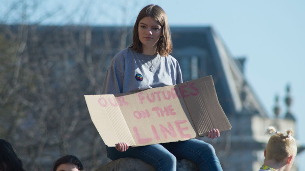 Photo taken in Leeds by Josh Barwick, used in event promotion, featuring a young woman in a gray t-shirt and jeans holding a cardboard sign emblazoned with the message, "Our Future's on the Line." The heads of three other young people are visible in the background, along with a more distant tree and building.