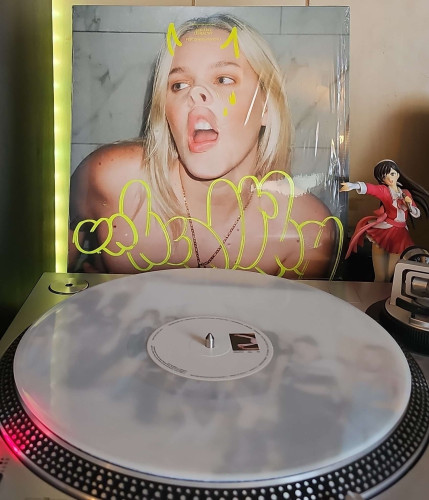 A milky clear vinyl record sits on a turntable. Behind the turntable, a vinyl album outer sleeve is displayed. The front cover shows Anne Marie pressing her lips and nose against glass. The plastic wrap is still on the cover and provides cartoon style horns and tears. 

To the right of the album cover is an anime figure of Yuki Morikawa singing in to a microphone and holding her arm out.