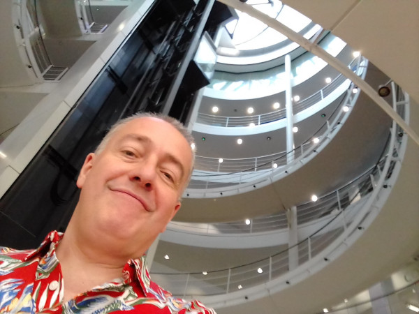 Colour photograph of myself in red patterned shirt standing on the spectacular spiral staircase at the heart of LSE Library.