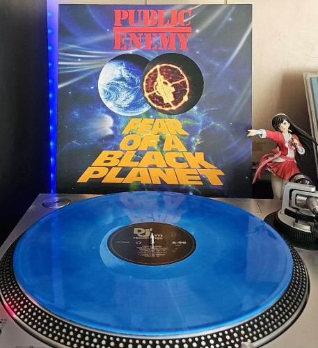 A Blue Swirl vinyl record sits on a turntable. Behind the turntable, a vinyl album outer sleeve is displayed. The front cover shows a galaxy with the planet Earth being collided with a planet showing the the Public Enemy symbol on it. 

To the right of the album cover is an anime figure of Yuki Morikawa singing in to a microphone and holding her arm out. 
