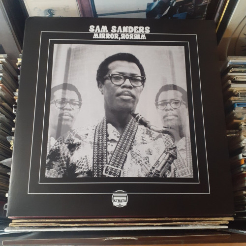 Album cover features a photo of Sam Sanders. He's wearing eyeglasses and has a saxophone in his hands,  and a wide strap for the saxophone around his neck.