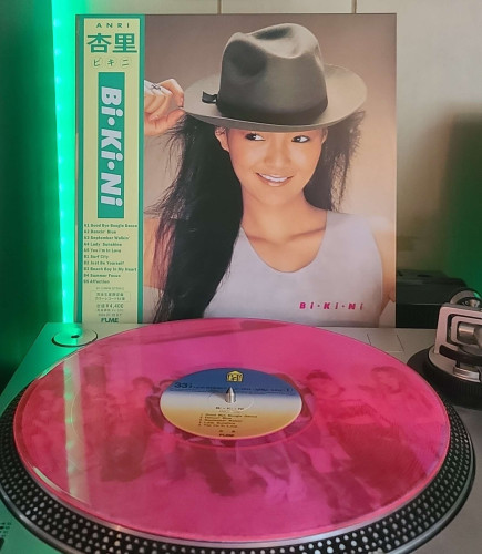 Image shows a turntable with a Transparent Pink vinyl record on the platter. Behind the turntable vinyl album outer sleeve is displayed. The front cover shows ANRI wearing a sleeveless top as she hols a hat she's wearing on her head. She is looking to the left away from the camera. 