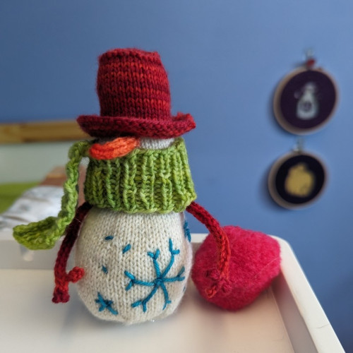 A knitted snowman in the style of Imagined Landscapes' gnomes (that is, hat tight to nose and no eyes). Here's wearing a top hat, has a carrot nose, a scarf, and embroidered snowflakes on his body.