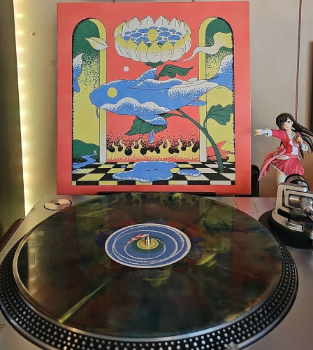A Rainbow Haze vinyl record sits on a turntable. Behind the turntable, a vinyl album outer sleeve is displayed. The front cover shows a koi fish swimming through a room with a large flower in it. 

To the right of the album cover is an anime figure of Yuki Morikawa singing in to a microphone and holding her arm out. 