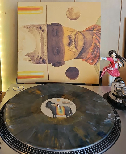 A Metallic Marble vinyl record sits on a turntable. Behind the turntable, a vinyl album outer sleeve is displayed. The front cover shows a man that is dressed like a cowboy.  There are moons to each side of him, and his hat is showing a desert like landscape. 

To the right of the album cover is an anime figure of Yuki Morikawa singing in to a microphone and holding her arm out. 