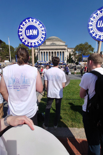 The Columbia library in the background, postdocs in white tshirts are holding UAW signs and looking away from the camera. The tshirts say "better working conditions for better research" on the backs we can see