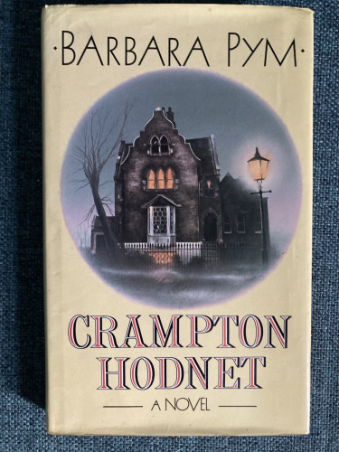 Cover of Crampton Hodnet by Barbara Pym. At the top of the cover is the author’s name in a Art Deco font. Beneath the author’s name is a circular illustration of a gothic looking four storey house, at dusk, with a lit window on the first floor and in the basement. Outside the house is an old streetlight which is illuminated, a tree with no leaves on it and some mist near the ground which gives it an eerie feel. Beneath the illustration is the book’s title ‘Crompton Hodnet - A Novel’.