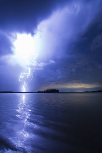 A powerful lightning strike forks out of dense, heavy low-hanging clouds. The scene is lit in blue from the lightning's illumination, with a faint gold streak along the horizon to the right from distant city glow. The lake below reflects back the sky and the path of the lightning, and along the horizon are silhouetted forests and a small, forested island dead centre.