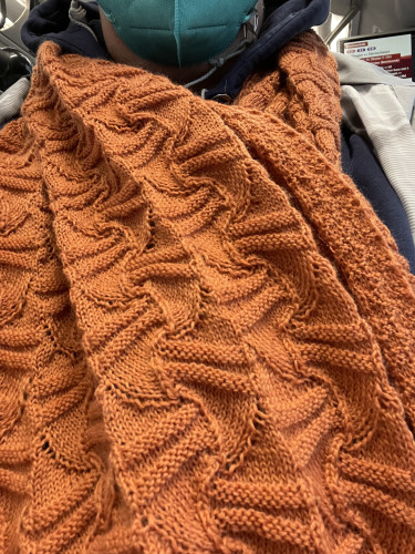Oranger Schal im Rechts-links-Muster mit einem kleinen Stück Hals sowie rechts und links hellgraue Jacke. 