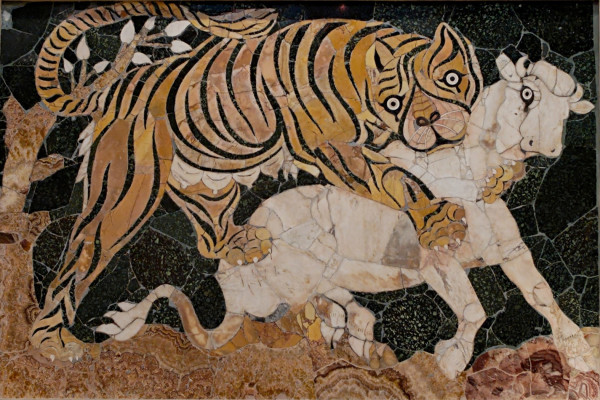 A tiger attacks a white calf. The tiger is shown in the moment of making contact with its front paws wrapped around the calf’s neck and chest. The calf has a wide eyed look as does the tiger. The background is dark green marble.
