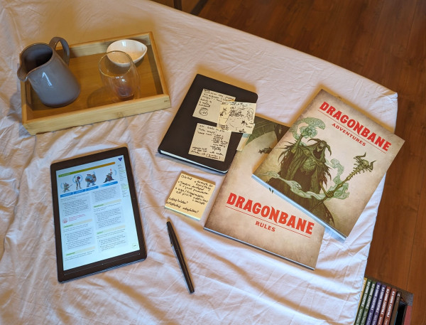 The corner of my unmade bed: displayed on a white sheet around with a small wooden tray with a jar and coffee glass, are two books from Dragonbane (red title, green illustration of a lich), a black notebook, with post-it notes stuck to it (ideas and a map for my BREAK!! game), the black felt pen I used, an my small tablet with the exploration rules for BREAK!!. 
The books of Old-School Essentials Basic and Advanced, stacked on the floor by the bed, are visible in a corner of the image.