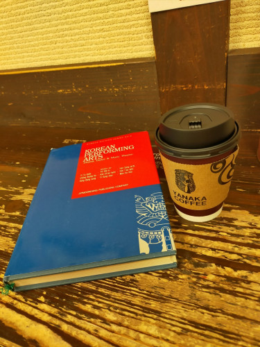 The photo is of a hardcover book. It is blue with a red square in the right corner featuring the title & authors. There's a design white illustration in the lower right of a tigerish image. To the right is a reddish brown coffee cup with a black lid & light brown coffee jacket of the Yanaka Coffee logo