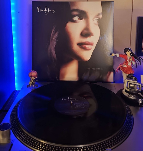 A black vinyl record sits on a turntable. Behind the turntable, a vinyl album outer sleeve is displayed. The front cover shows Norah Jones looking off to her side. 