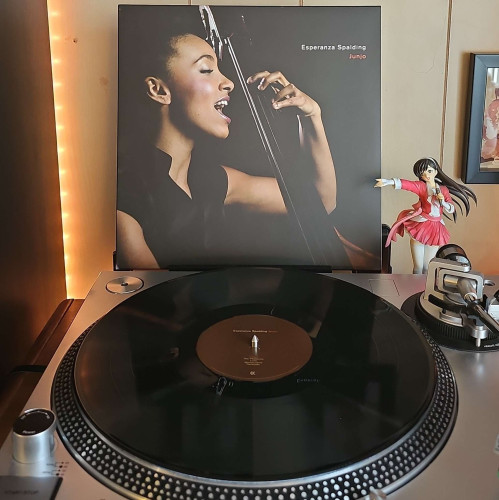 A black vinyl record sits on a turntable. Behind the turntable, a vinyl album outer sleeve is displayed. The front cover shows Esperanza Spalding playing a bass and singing. 