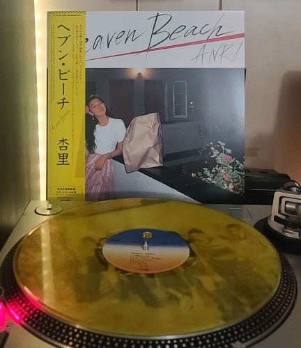 Image shows a turntable with a Transparent Yellow vinyl record on the platter. Behind the turntable vinyl album outer sleeve is displayed. The front cover shows ANRI leaning against the top of a railing, with a paper bag next to her. There is a pair of doors and windows behind her. 