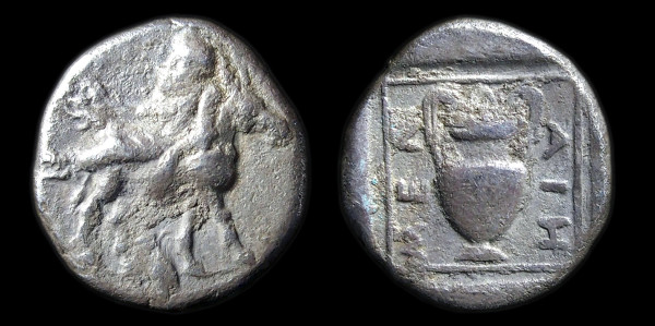 Small silver coin, obverse and reverse, shown against black background. On the obverse, a drunken man, holding a kantharos, lying on the back of a donkey/ass. On the reverse, an amphora surrounded by the letters MENΔΑΙΗ