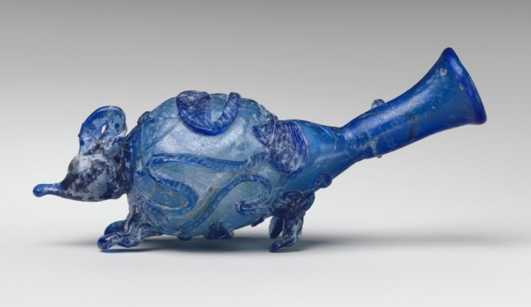 Description from the museum: “Translucent cobalt blue, with same color added head, ears, eyes, feet, and snake-thread decoration.
Rounded and thickened rim; funnel-shaped, elongated neck with tooling marks around base; misshapen piriform body, with separate large blob of glass (solid?) applied to rounded bottom of body.
The neck has been turned upwards to appear like the mouse’s tail; added to the body of the vessel to create the animal are four separate tooled trails for the feet and for the head a large blob that has been drawn out to make a pointed nose; ears and eyes have also been added to the head; the body is further decorated with snake-thread trails, all flattened and notched, in the form of two long-necked, thin-legged birds, one on the animal’s back, the other on his belly between his feet, interspersed with foliage comprising tendrils and ivy leaves; a plain spiral trail is wound twice around the lower part of the neck.”