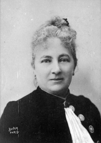 A black and white photograph of Juliette Adams from about 1895. She looks directly at the viewer, her light curly hair pulled up and back. She wears a high-necked dress with large medallion buttons.