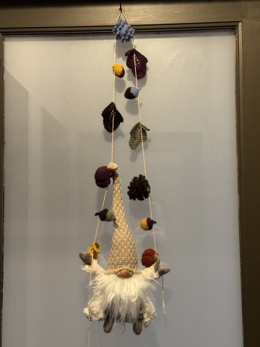 Gnome hanging from door. Gnomes hands grip a string decorated with autumn knitted items (acorns, leaves, pumpkins)