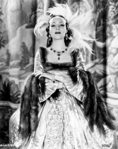 A photo of Dolores del Rio in Madame du Barry (1934). It is in black and white and features a confident looking woman looking straight at the viewer. She wears shiny a shiny costume historically inspired gown lined in furs. 