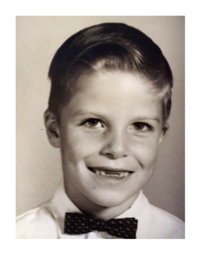 1960s school picture. white kid with slicked back, short hair wearing a white shirt and clip-on bowtie. missing his two front teeth and making eye contact, smiling.