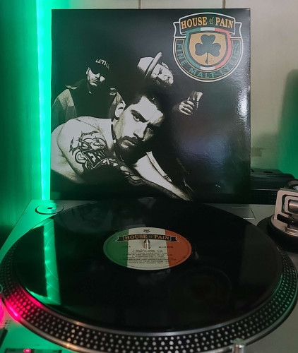 Image shows a turntable with a black vinyl record on the platter. Behind the turntable vinyl album outer sleeve is displayed. The front cover shows 3 members of House of Pain mean mugging the camera. 