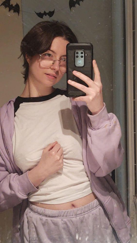 selfie of poki (slim white person with short dark hair) taken in a mirror. They're juste there being cute, wearing their prescription glasses, an oversized purple hoodie, purple jersey shorts and a white and black raglan tee