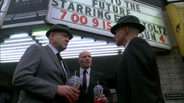 Three pale, hairless men in plain grey suits standing in front of a movie theatre entrance, with the board above it advertising "Back to the Future, starring Eric Stoltz".