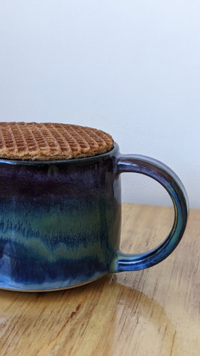 A picture of a mug, half in shot, upon which sits a stroopwafel. The mug is dark but swirled with greens and blues horizontally. If stands on a light wooden desk with a simple white wall as the background