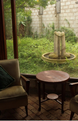 Foto von einem Sitzplatz vor einem großen Fenster mit 2 Sesseln und einem Tisch auf steinernem Fußboden. Das Fenster ist mit Blick auf eine große Schale mit 3 großen Phallen innerhalb einer Grünfläche, dahinter eine Steinmauer. 