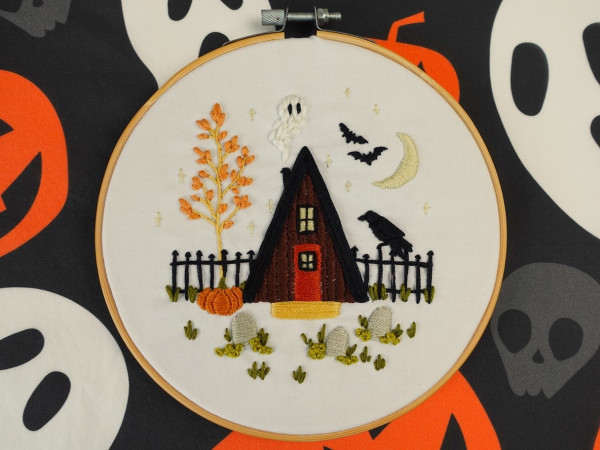 Hand embroidery piece finished in a wooden hoop. It's a wooden cabin surrounded by an autumn tree, pumpkin, gravestones, and a raven with bats in the sky and a ghost appearing from the chimney. It's clearly beginner work but is cute anyway!