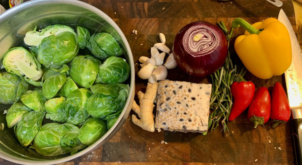A cutting board on which sits a steel bowl of halved brussels sprouts in water, a piced of ginger root, about 10 garlic cloves, a block of tempeh (white, with dark spots where black beans show through) a red onion, a small pile of fresh rosemary, 3 red Fresno peppers, a large yellow bell pepper, and a Japanese chef's knife.