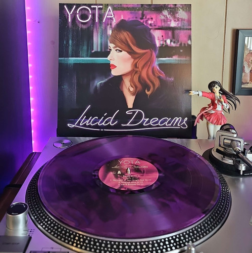 A Purple Transparent vinyl record sits on a turntable. Behind the turntable, a vinyl album outer sleeve is displayed. The front cover shows a painting of Yota's side profile . 