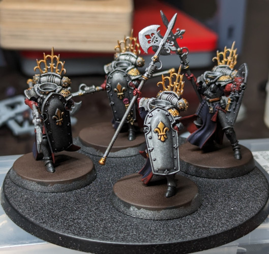 Warhammer 40k Adepta Sororitas Celestian Sacresants armed with anointed halberds. Metallic black armor with gold accents and purple robes with red trim. No one has their heads on, but I finished all the heads earlier, which makes things much easier now.