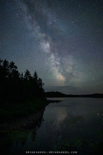 The Milky Way rises vertically through a deep blue dark sky. Below the Milky Way there's a peninsula covered with a boreal forest. Both the Boreal forest and the Milky Way reflect into a completely calm lake.