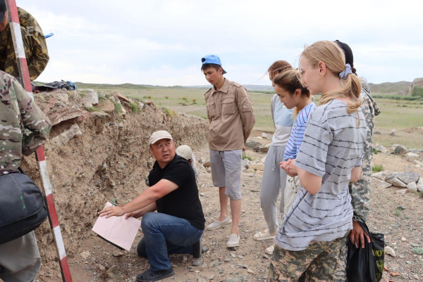 Setting: mostly flat grassy steppe landscape. Foreground: group of young students standing, listening to an older male instructor holding a book and kneeling in front of a mound. He is gesturing towards the mound and speaking to the students.