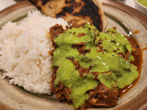 A bowl with steamed white rice, a mound of rich red curry and a piece of grilled paratha. The curry has a bright green sauce drizzled over it.