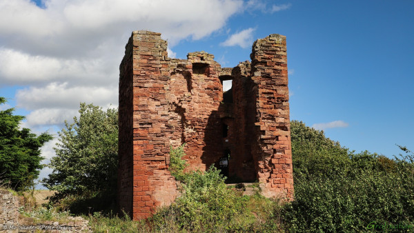 The scene is of a red brick, ruined castle surrounded by green trees and shrubs. The sky is blue with some white cloud and the sunlight is from above right. The building is a shell, in places the outer walls have completely fallen, and what was the interior can be seen clearly. The brickwork itself is rotted and crumbling but the outer walls still hold the general shape of a rectangular block building.