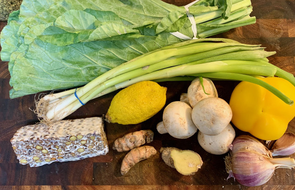a large bunch of collard greens, a   bunch of scallions a block of tempeh, a lemon, four mushrooms, two turmericc roots a piece of ginger root a yellow bell pepper, and a large head of red garlic.