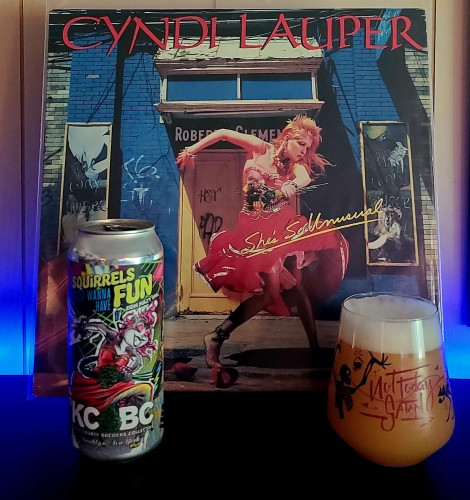 A stemless glass filled with beer and a 16oz can sit on a shelf. The glass features a skeleton and a skeleton cat, and says "Not Today Satan". The can artwork shows a squirrel dressed as Cyndi Lauper doing the iconic pose from the "She's So Unusual" album. Instead of holding a bouquet of flowers, the squirrel is holding a bouquet of hops. 

Behind the glass and can is the actual vinyl album of Cyndi Lauper's "She's So Unusual" on a display stand. 