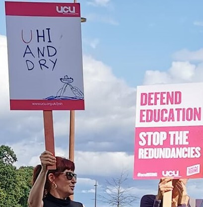 me holding up a plackard with UCU branding which says UHI AND DRY with a cartoon of a small boat stranded on dry land. Next to me someone is holding a plackard which says Defend Education: Stop the redundancies