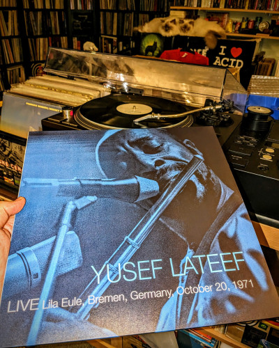 in the front a hand holding the blue LP cover of "Yusef Lateef - Live Lila Eule, Bremen, Germany, October 20, 1971 with a Yusef headshot, eyes closed, playing the flute into two microphones. behind it the vinyl is playing on the technics. in the background a record collection wall, also two cats Emil & Milo cuddling on the shelf.
