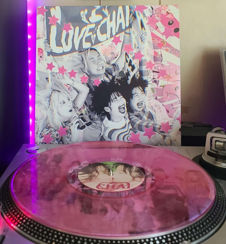 A translucent pink vinyl record sits on a turntable. Behind the turntable, a vinyl album outer sleeve is displayed. The front cover shows the 4 members of CHAI reacting to something off camera. 1 Is cheese grinning holding up a LOVE CHAI banner, another is holding binoculars and looking dumbfounded. The other two are happily yelling with their eyes closed.
