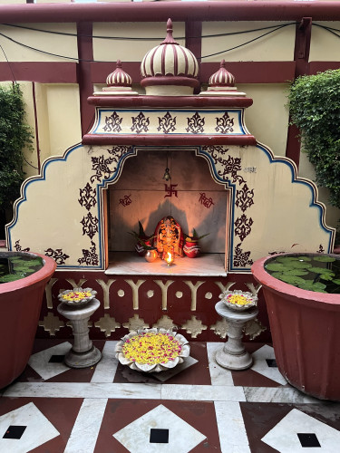An altar to Ganesh in a small stone alcove at our hotel in Rajasthan. The alcove is made of brick and is painted off white with ochre red and blue trimmings. The idol is inside a small alcove decorated and with a light lit in front of it. The whole setup has two large clay pots with live lotuses floating in water and three marble bowls with flower petals inside.  