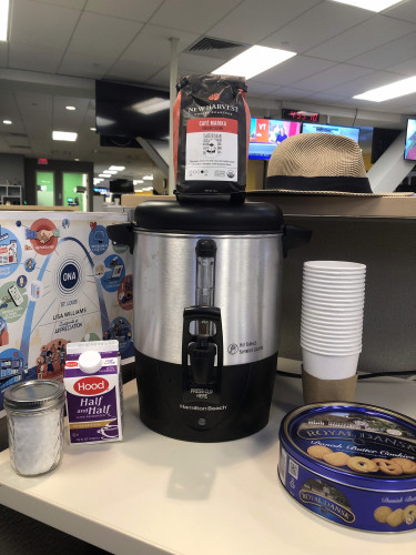 A large percolator sits on a desk in an open newsroom. Beside the percolator are paper cups, a pint of half and half, a jar of sugar, and a tin of butter cookies. A bag of coffee sits on top of the percolator. Behind can be seen a straw hat on a shelf, and further in the background, TV screens and a larger office can be seen. 