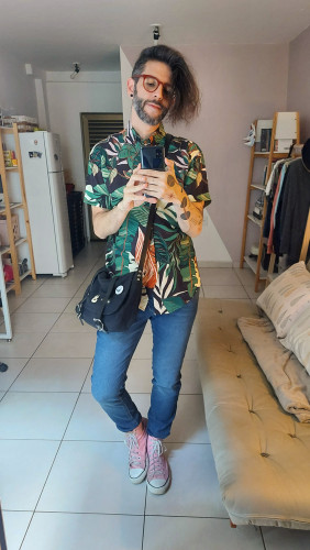 flux taking selfie in front of a mirror, showing off his look which includes a pink converse, blue jeans, a buttoned up shirt with a foliage print and a black shoulder bag with some pins attached to it.