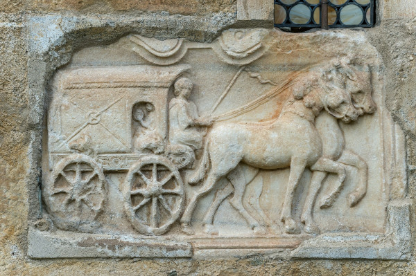 Relief of a carriage with two horses. The carriage has a small opening to see an occupant while the driver sits out front with reins and a switch.