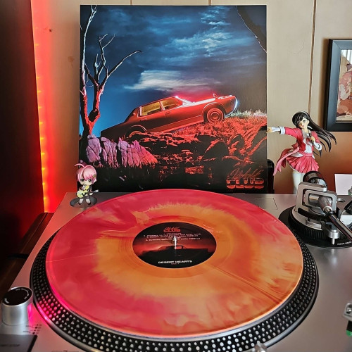A CRASH (Pink, red, orange, yellow sunburst)vinyl record sits on a turntable. Behind the turntable, a vinyl album outer sleeve is displayed. The front cover shows a classic car on a hill in the desert. Rocks and barren trees are in the foreground of the image, and a dark blue sky in the background. 