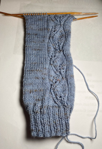The hand-knitted leg of a sock with a lace pattern that resembles a vine
