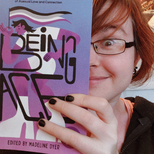 A red-headed white nerd peeking out from behind a purple and blue book called BEING ACE, they have glasses and black nails and look a bit evilly gleeful to be published in such an amazing volume! 